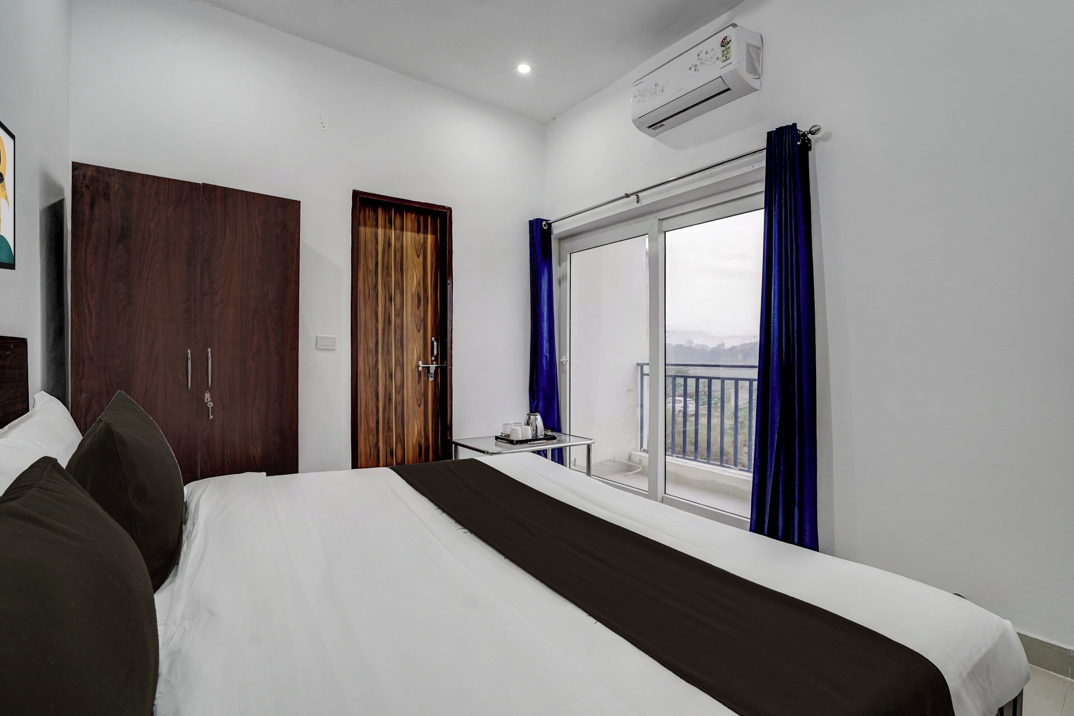 Una habitación de hotel con una cama, un armario, una mesita con una bandeja y una ventana con cortinas.