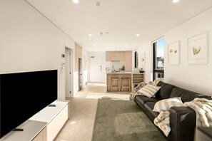 A modern living room with a flat-screen TV, a leather sofa, and a kitchen in the background. - Beautifully Furnished 1-Bed in the City Centre (Christchurch)
