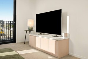 Ein modernes Wohnzimmer mit einem großen Flachbildfernseher auf einem weißen Schrank, einem kleinen Beistelltisch mit Lampe und Blick auf eine Baustelle durch eine Glastür.