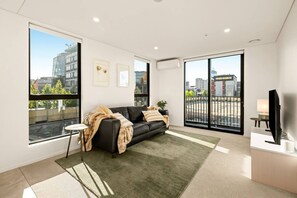 A modern living room with a black leather sofa, a small side table, a flat-screen TV, and large windows offering a city view. - Beautifully Furnished 1-Bed in the City Centre (Christchurch)