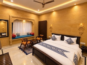 Quarto de hotel com uma cama grande, área de estar com sofá e poltronas, ventilador de teto e arandelas decorativas.