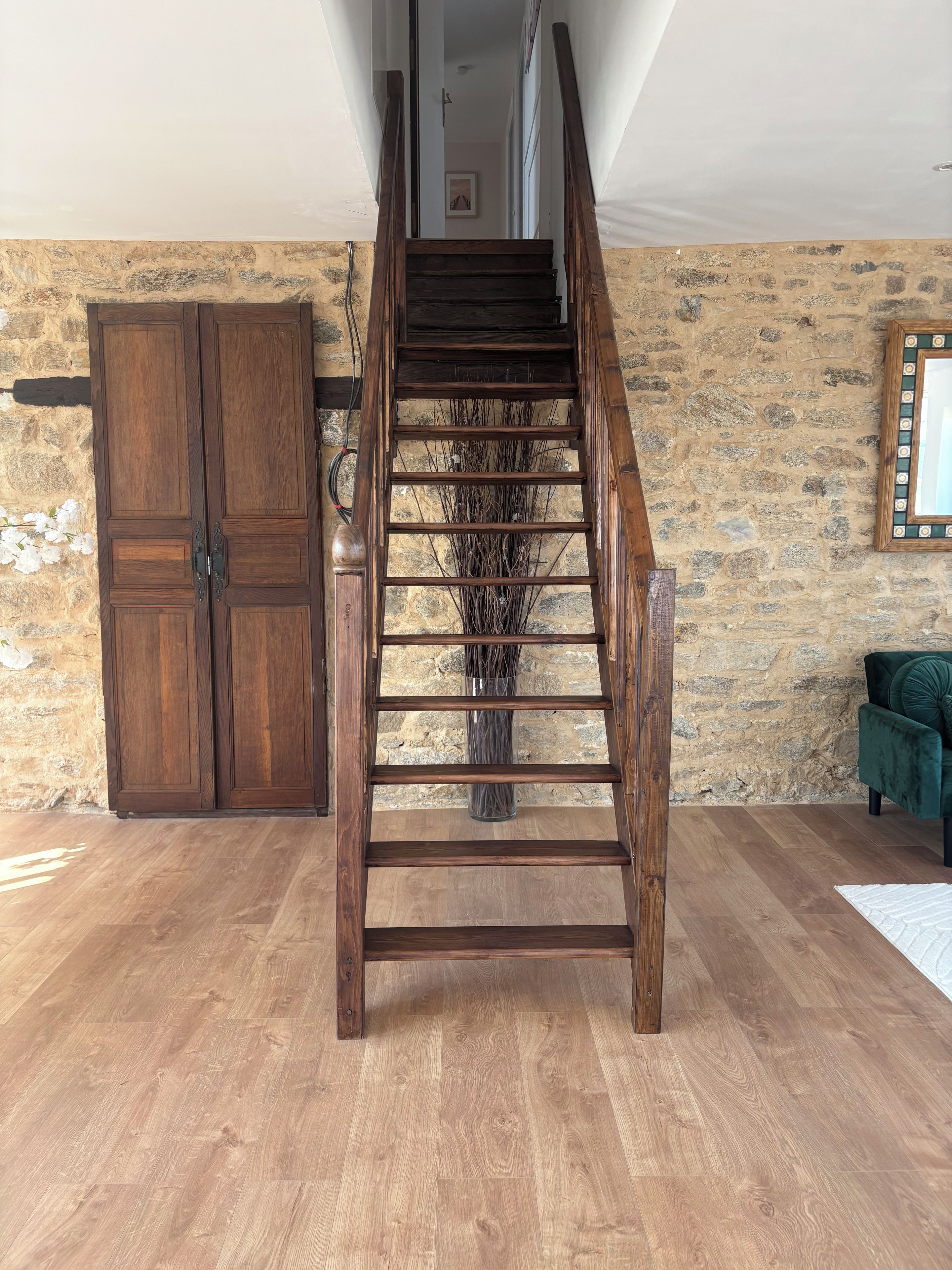 A wooden staircase leading to a loft area with a stone wall and a green upholstered chair.