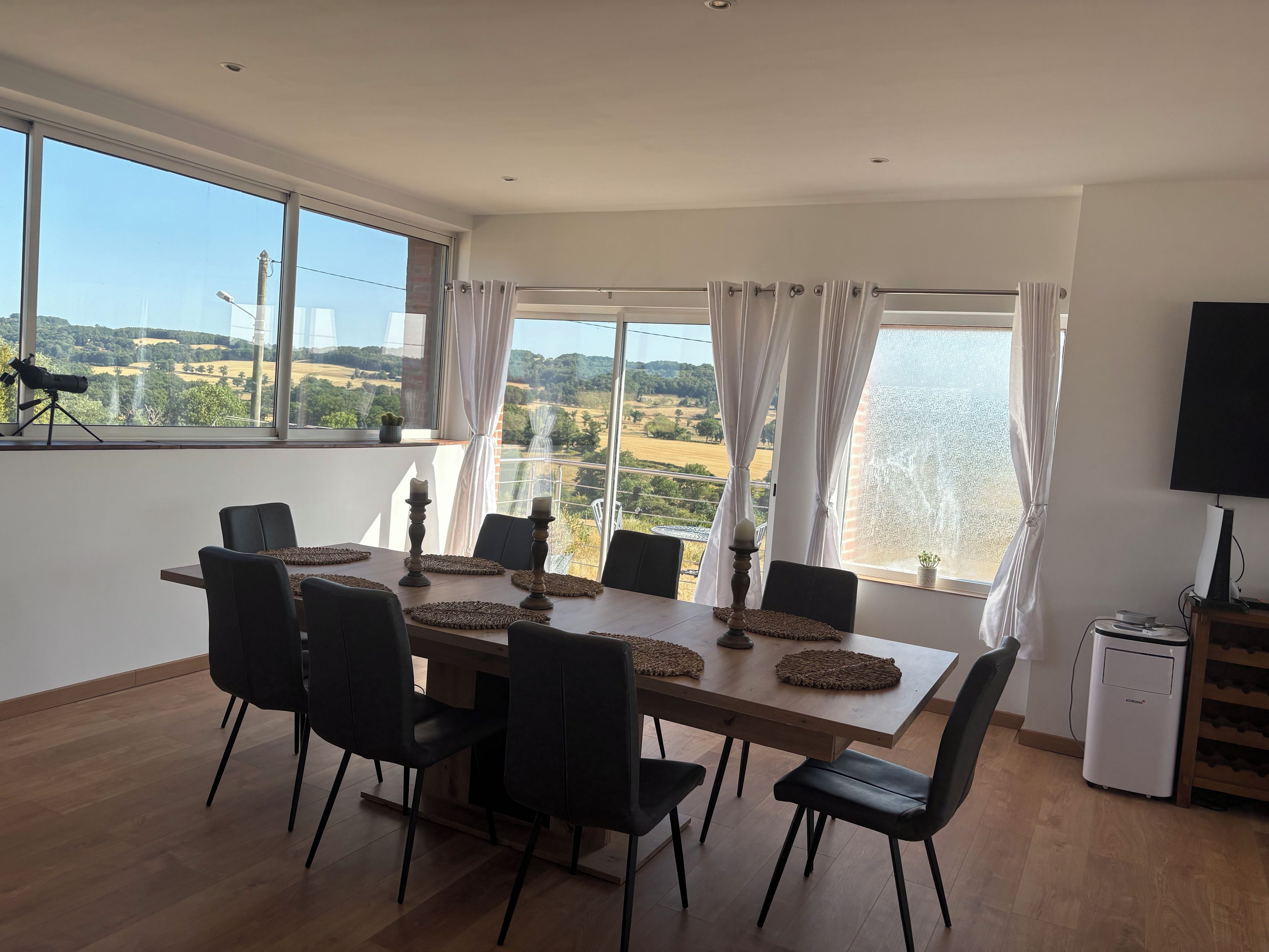 Un espace repas comprenant une grande table, des chaises et offrant une vue sur un paysage grâce à de larges fenêtres.
