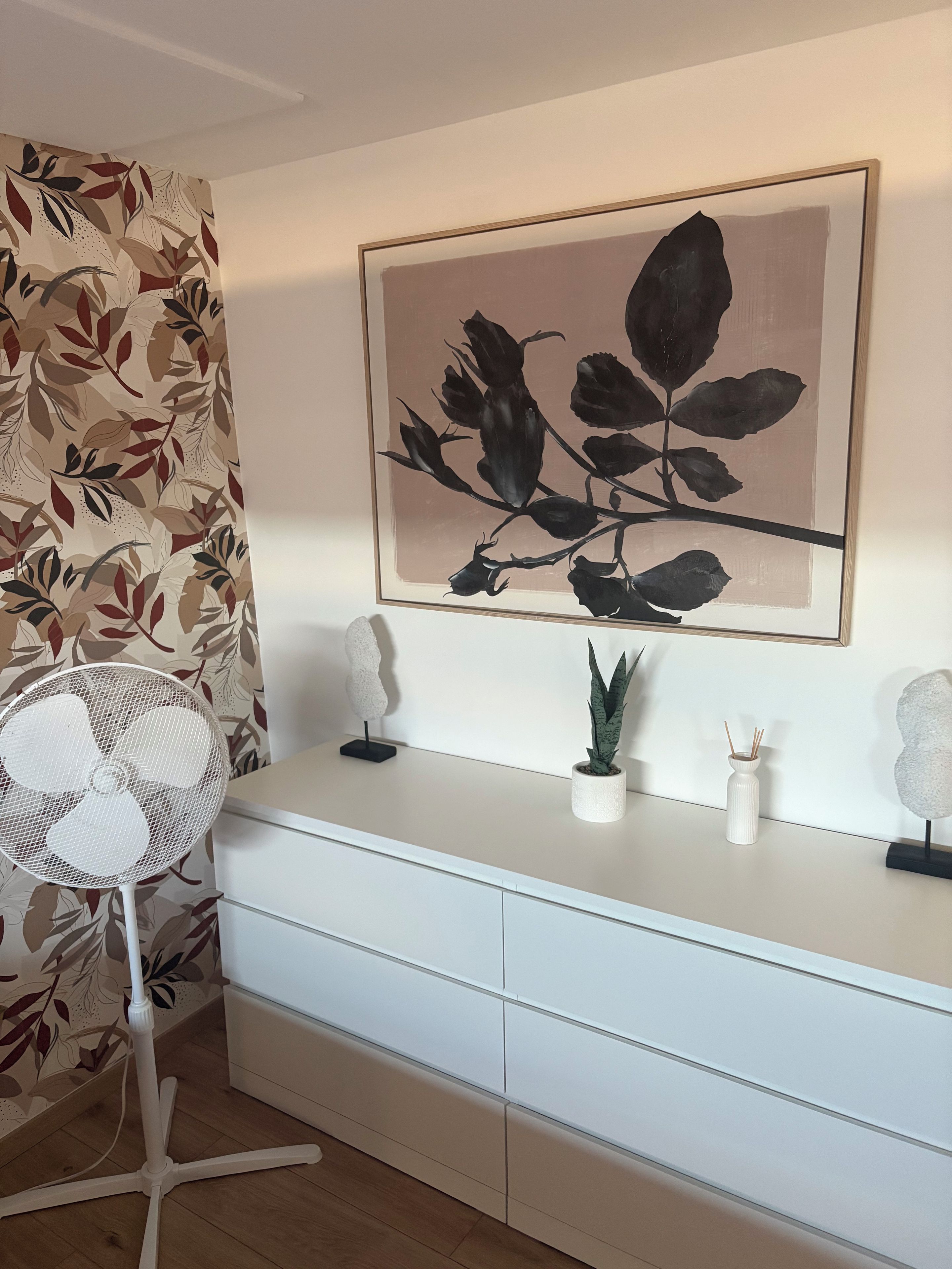 A room with a white desk, a framed botanical print, a white fan, and a potted plant.