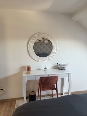 Ett sovrum med en säng, ett vitt skrivbord med en spegel, en röd stol och ett litet runt bord med diverse föremål på.