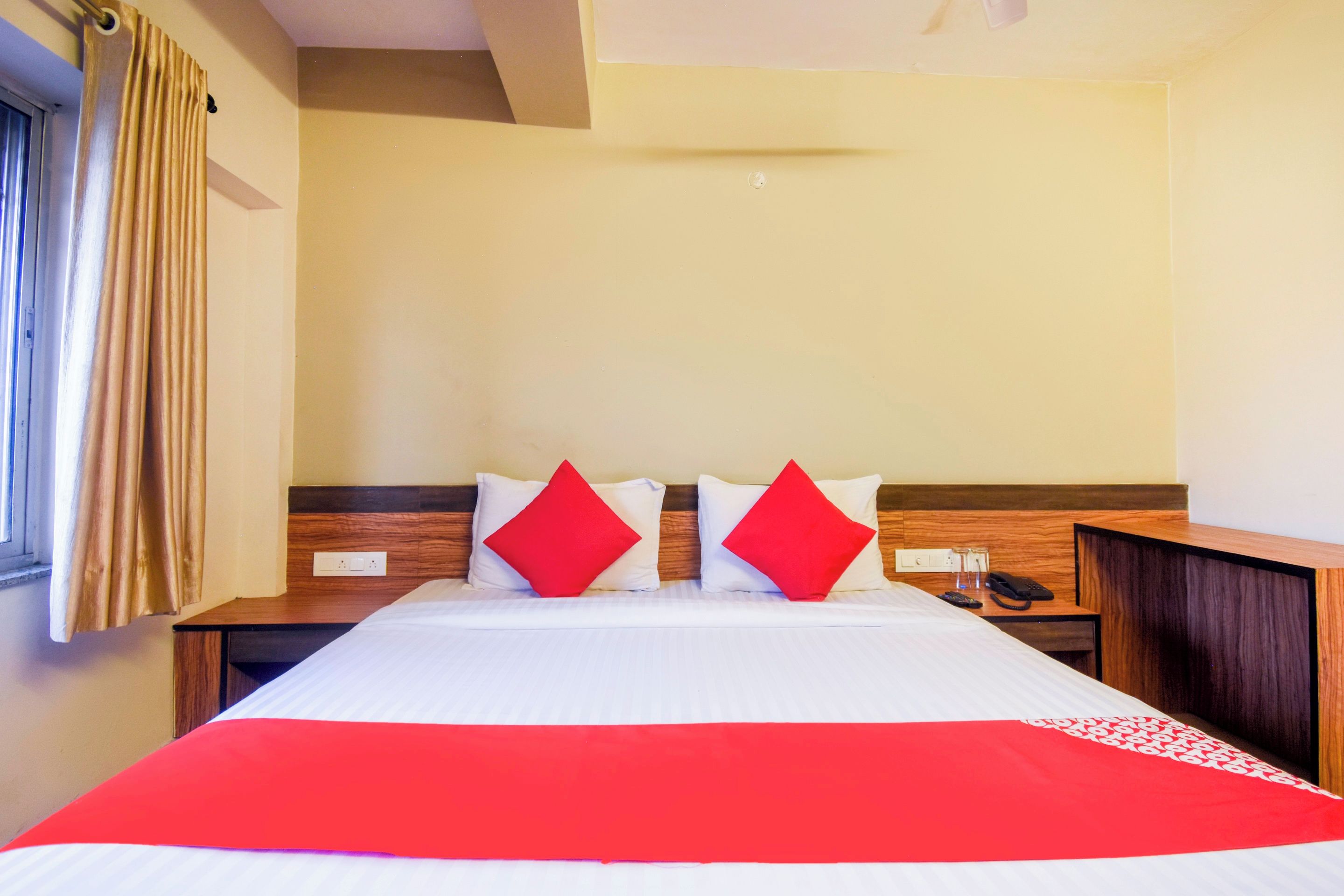 A hotel room with a bed, two nightstands, a wooden headboard, a red and white bedspread, and a window with curtains.