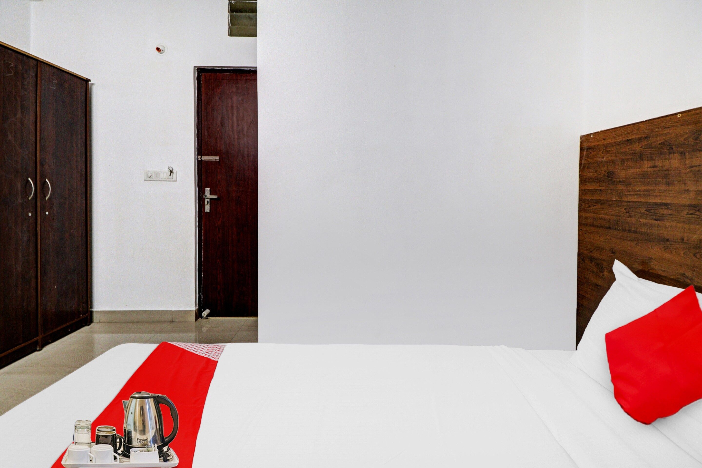 A hotel room with a bed, a wooden wardrobe, a door, and a bedside table with a kettle and cups.