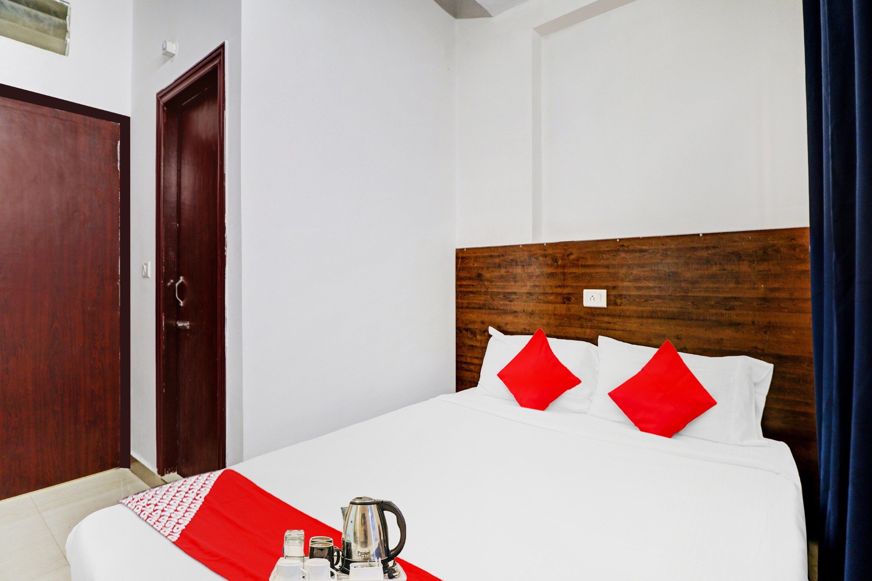A hotel room with a wooden headboard, white bedding, red pillows, and a breakfast tray with a kettle.