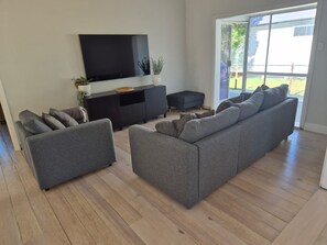 A modern living room with a flat-screen TV, grey sofas, a black entertainment unit, and a sliding glass door leading to a balcony. - Breeze on Prince: Spacious cottage offering modern comfort (Waratah)