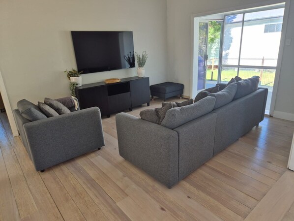 A modern living room with a flat-screen TV, grey sofas, a black entertainment unit, and a sliding glass door leading to a balcony. - Breeze on Prince: Spacious cottage offering modern comfort (Waratah)