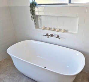 A white freestanding bathtub in a bathroom with white tiled walls and a window above. - Spacious home 'Breeze on Prince', offers modern connectivity and luxury comfort. (Waratah)