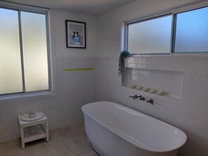 A bathroom with a freestanding bathtub, a small stool, a framed picture on the wall, and a window with frosted glass. - Breeze on Prince: Spacious cottage offering modern comfort (Waratah)