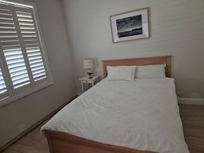 A bedroom with a bed, a nightstand, a lamp, a framed picture on the wall, and a window with blinds. - Breeze on Prince: Spacious cottage offering modern comfort (Waratah)