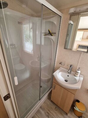 A bathroom with a glass-enclosed shower, a white sink, and a wooden cabinet. - Greenacres Holiday Park - Holiday Accommodation 20263 (Porthmadog)