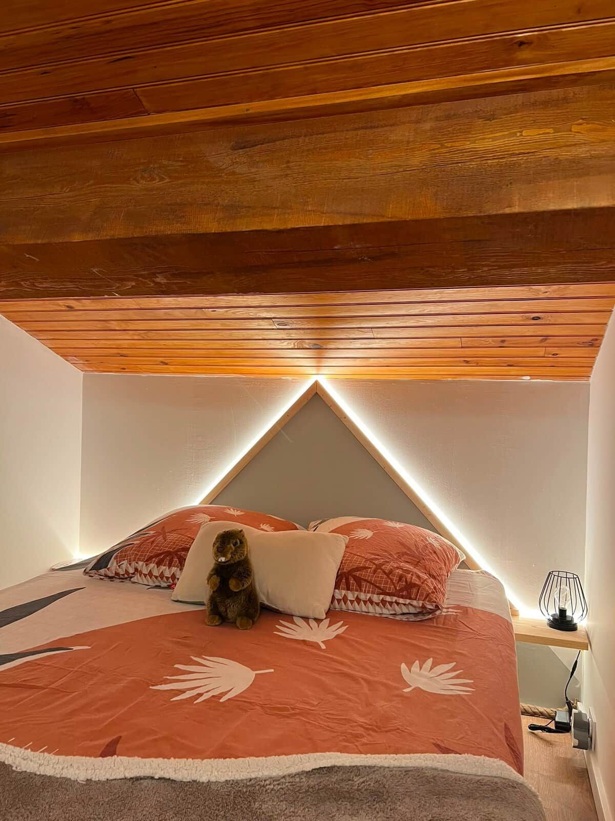 A bedroom with a sloped ceiling, a bed with a patterned bedspread, a teddy bear, and a bedside lamp.