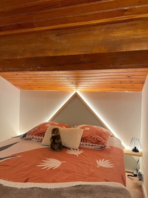 A bedroom with a sloped ceiling, a bed with a patterned bedspread, a teddy bear, and a bedside lamp.
