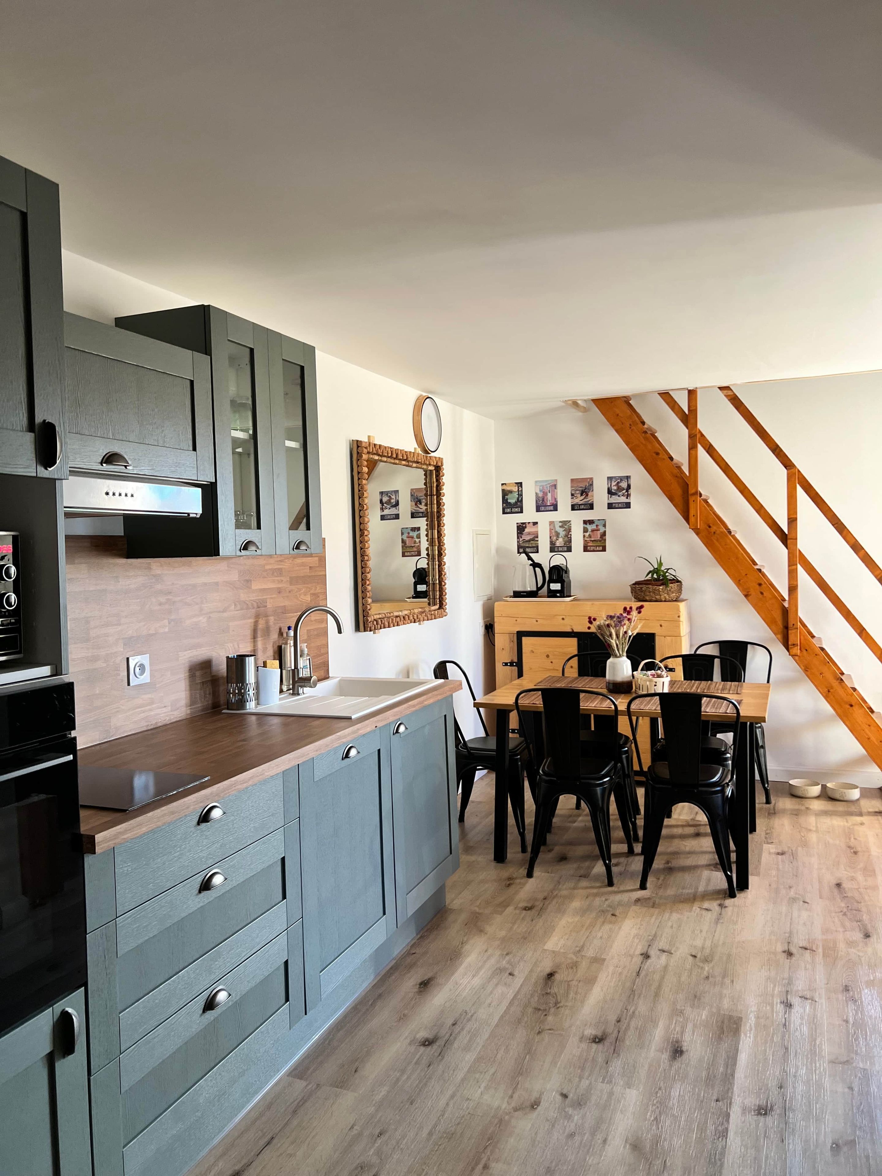 A modern kitchen with grey cabinets, a wooden dining table, and a staircase.