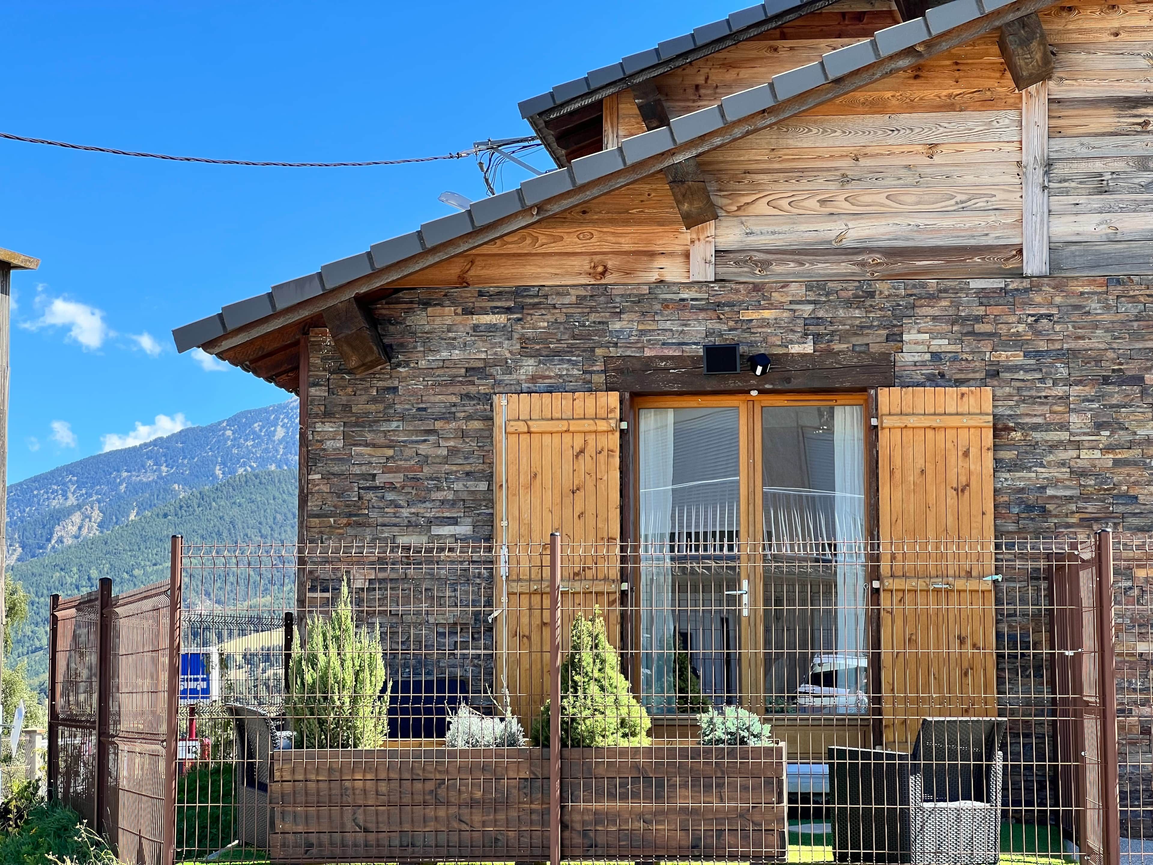 A wooden cabin with a stone facade, wooden shutters, and a fenced garden area.