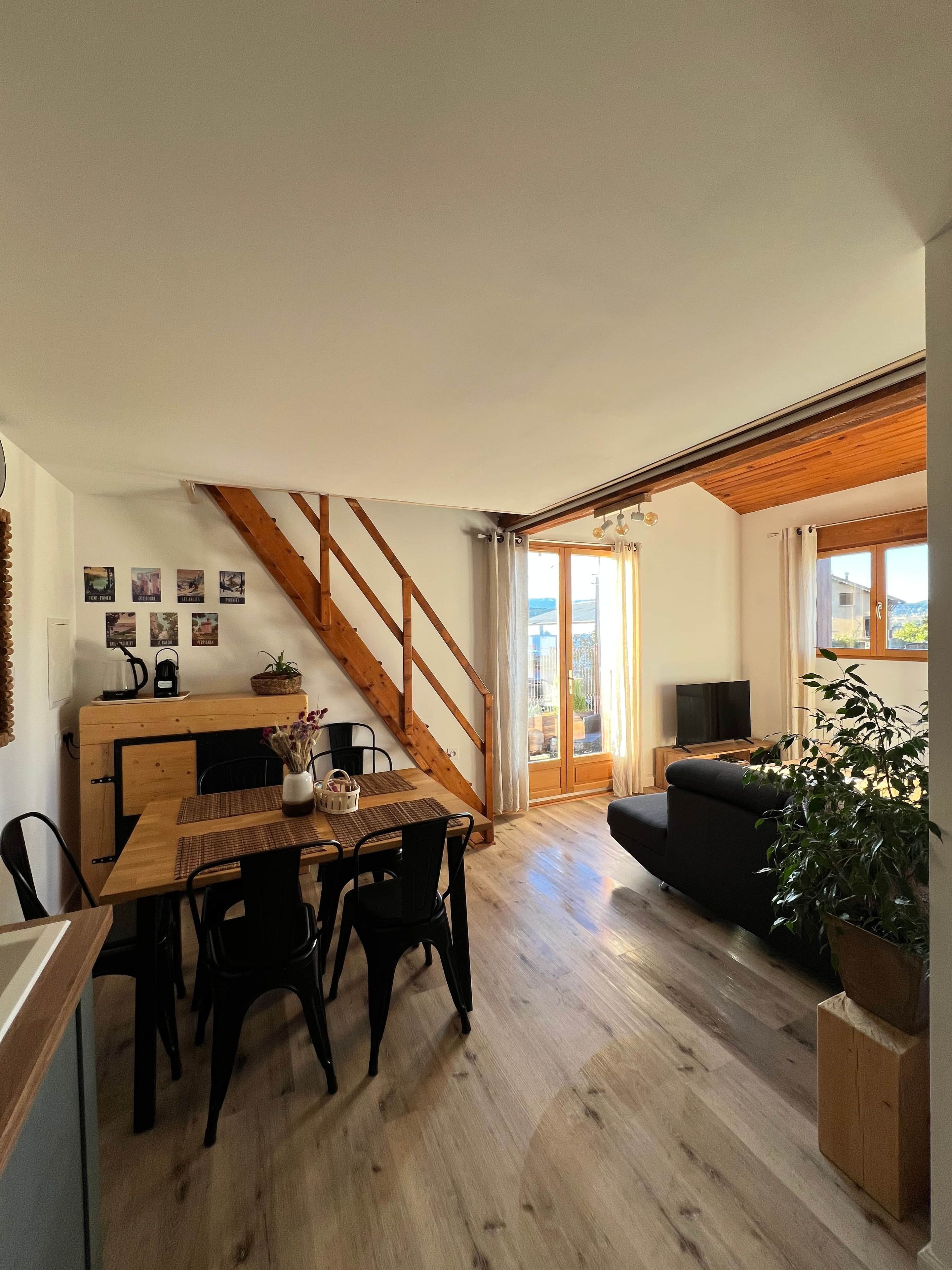 A spacious living area with a wooden staircase, a dining table with chairs, a sofa, and a large window with curtains.