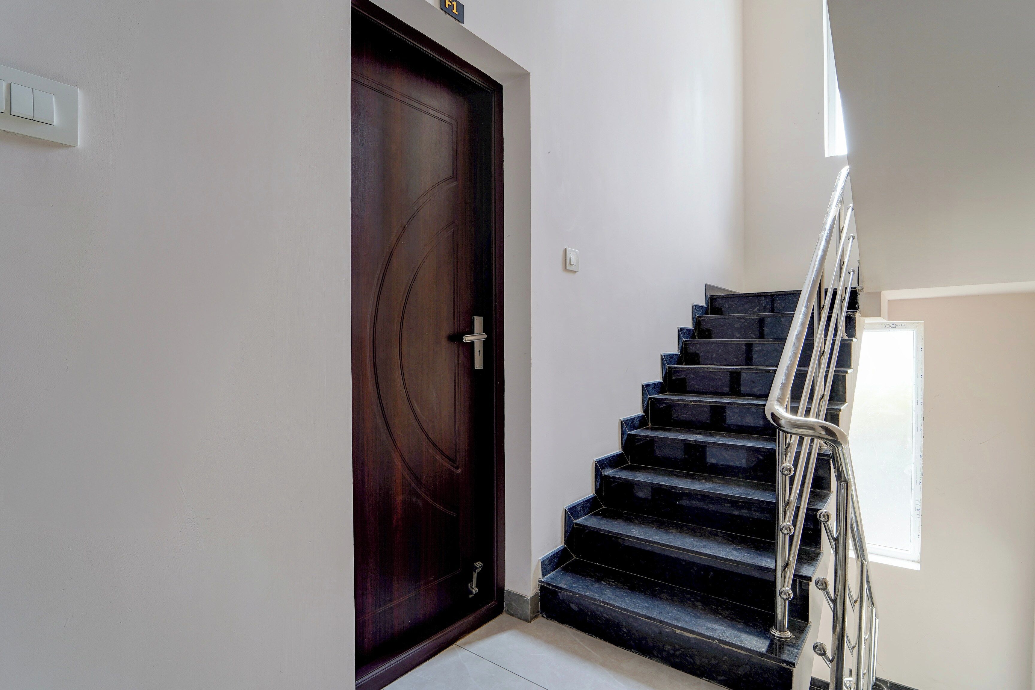 Una puerta de madera oscura con un picaporte, una pared blanca y una escalera con escalones negros y una barandilla de metal.