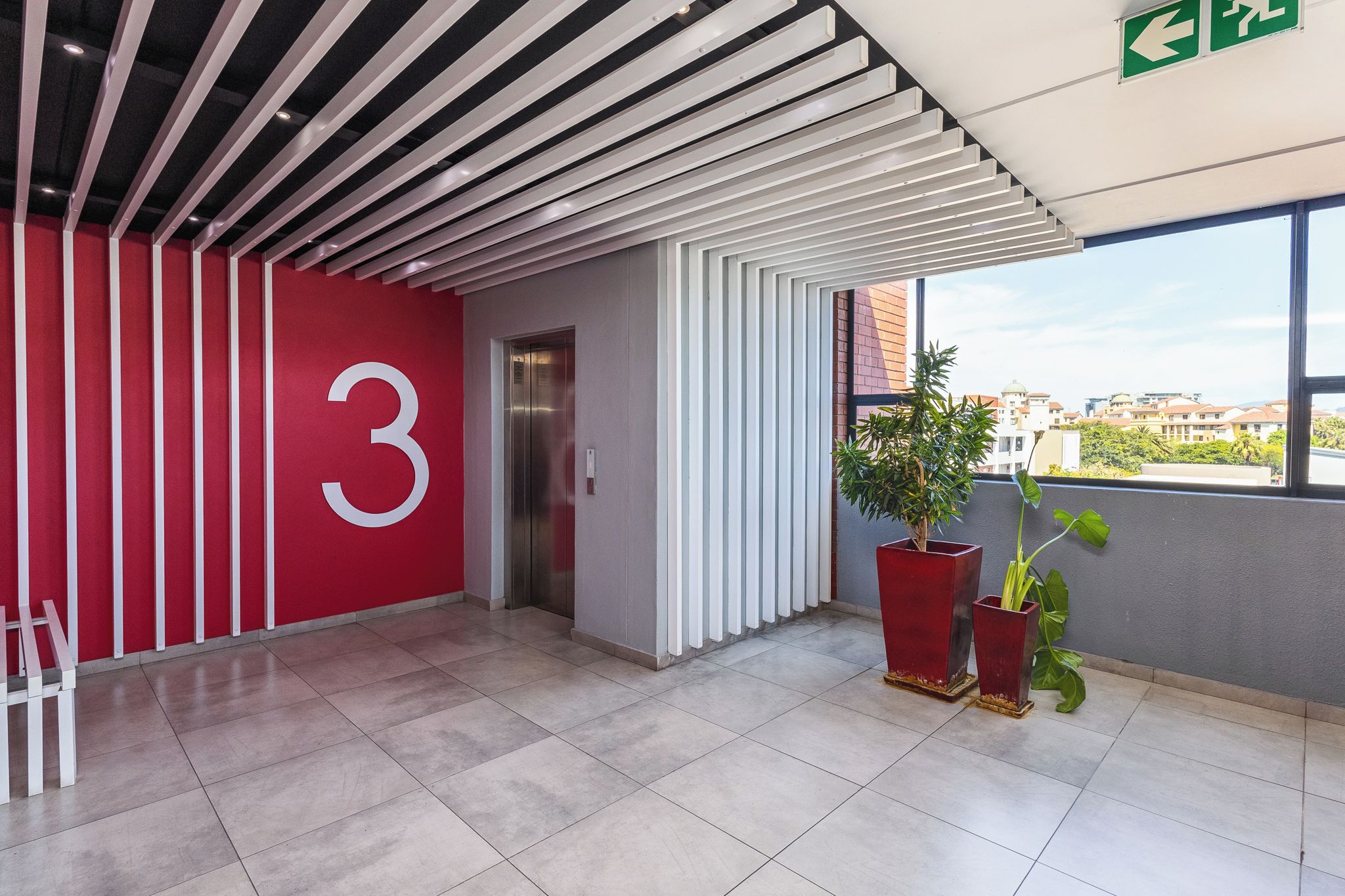 A modern interior with a red wall featuring a large number '3', a ceiling with horizontal beams, and a balcony with potted plants.