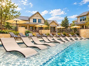 A poolside area with lounge chairs and umbrellas, a clear blue pool, and a building with a stone facade and multiple windows. - Designer Texan Stay - Resort Pool - Arcade & Games (Round Rock)
