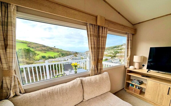 A room with a sofa, a television, and a view of a green landscape through large windows.