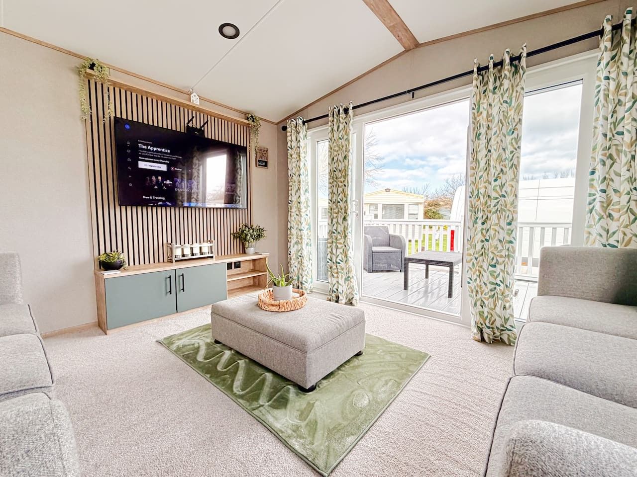 A modern living room with a flat-screen TV, a grey sofa, a matching ottoman, and a balcony with patterned curtains.