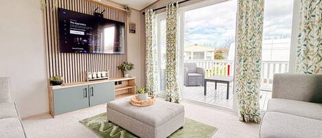 A modern living room with a flat-screen TV, a grey sofa, a matching ottoman, and a balcony with patterned curtains.