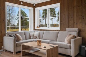A modern living room with a grey sofa, a wooden coffee table, and large windows offering a view of trees. - GV19– Family-Friendly Cabin at Trysilfjellet Arena (Trysil)