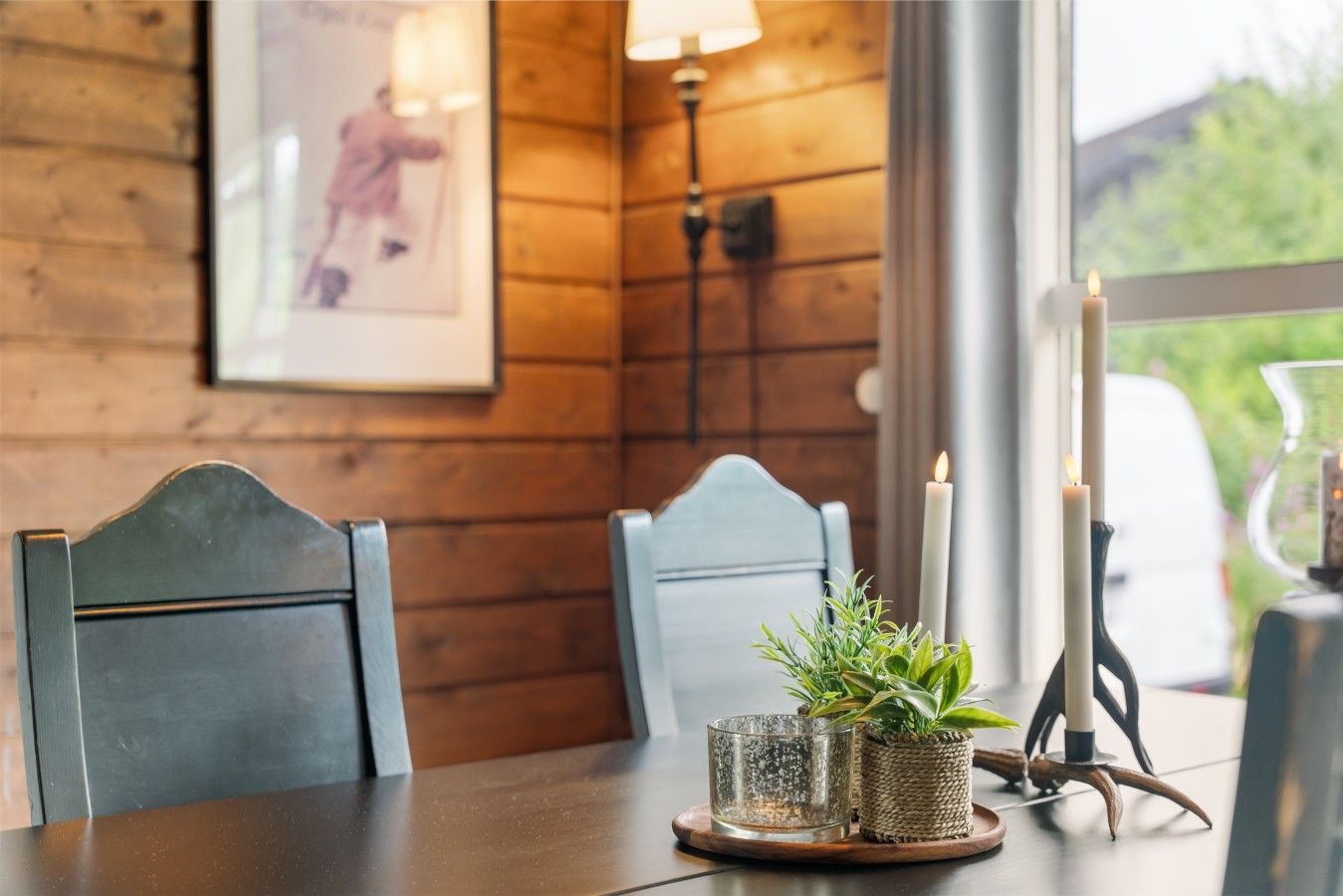 A wooden interior with a dining table, two chairs, a candle holder with candles, a glass candle holder with a plant, and a framed picture on the wall.