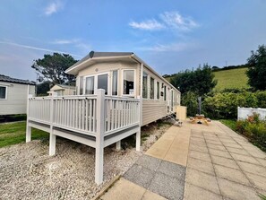 A single-story mobile home with a deck, surrounded by gravel and a paved walkway.