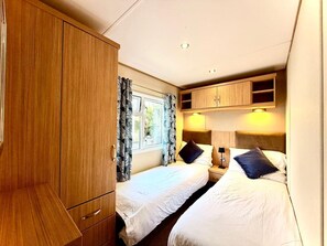 A compact bedroom with two beds, wooden wardrobe, and a window with curtains. - The Windermere - Luxury Retreat by the Sea in Devon (Challaborough)