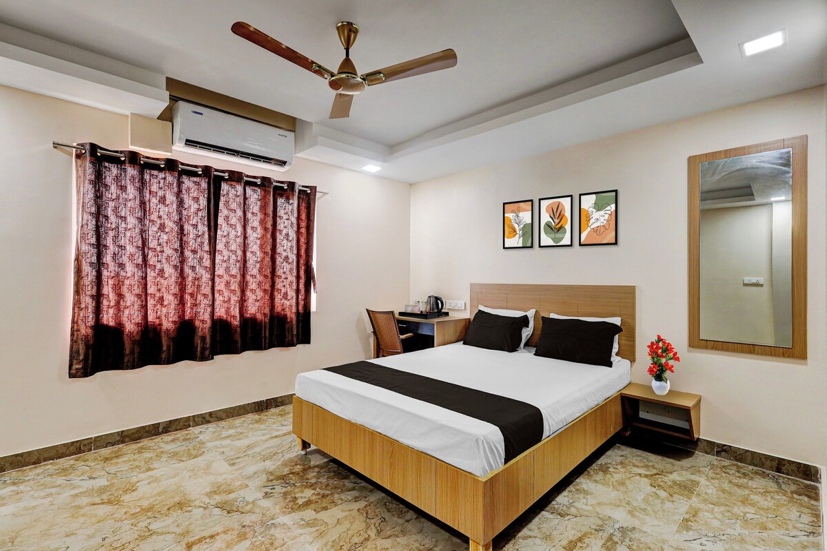 A hotel room with a bed, a nightstand, a desk, an air conditioning unit, a ceiling fan, a window with curtains, and wall art.