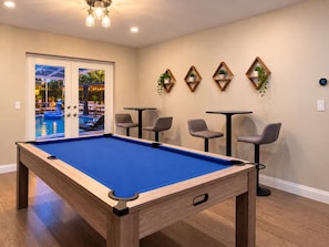 A pool table with a blue felt surface, surrounded by six grey chairs and two small round tables.