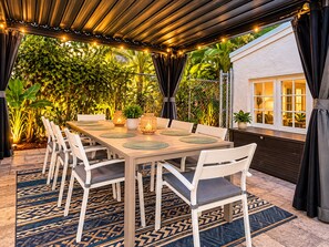 Un área de comedor al aire libre con una mesa dispuesta para seis personas, rodeada de sillas, bajo un toldo con luces colgantes.