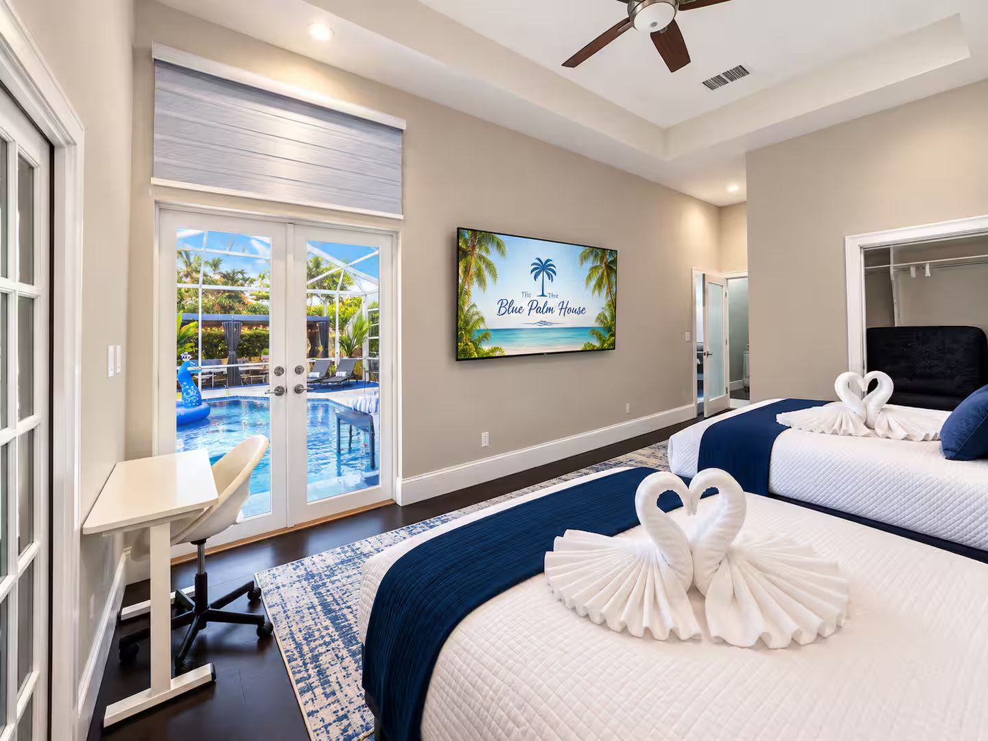 Habitación de hotel con vista a la piscina, dos camas, un escritorio y un televisor que muestra 'The Blue Palm House'.