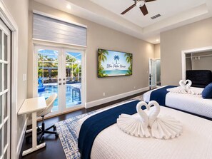 Habitación de hotel con vista a la piscina, dos camas, un escritorio y un televisor que muestra 'The Blue Palm House'.