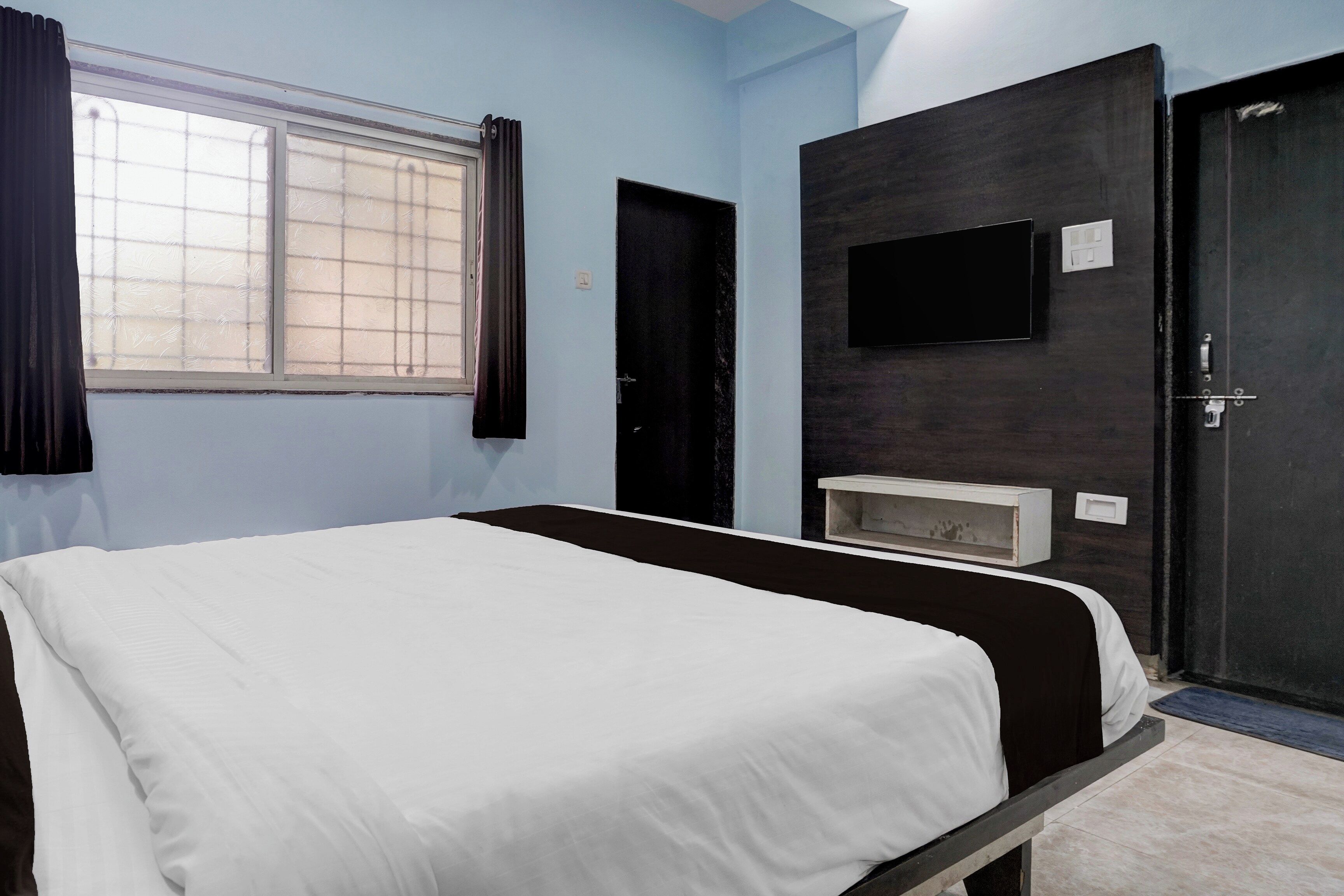 A room with a bed, a television mounted on the wall, a door, and a window with curtains.