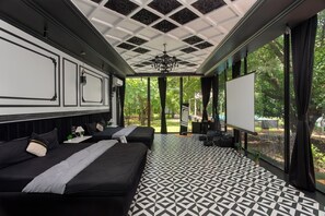 A hotel room with two beds, a projector screen, and a large window with a view of greenery.