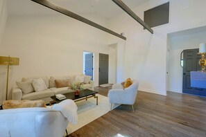 Ein geräumiges Wohnzimmer mit einer Couch, Sesseln, einem Couchtisch und einer Lampe. Holzdielen und eine Tür zu einem anderen Raum.