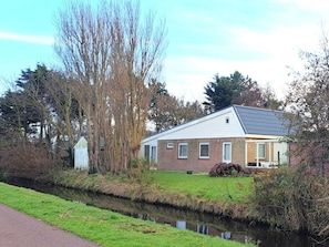 A single-story house with a sloped roof, a small front porch, and a narrow canal in front. - Bungalow With 3 Bedrooms (Julianadorp)