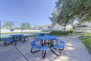 Des tables de pique-nique en plein air avec des bancs, un terrain de sport clôturé et, au fond, des immeubles d’habitation.