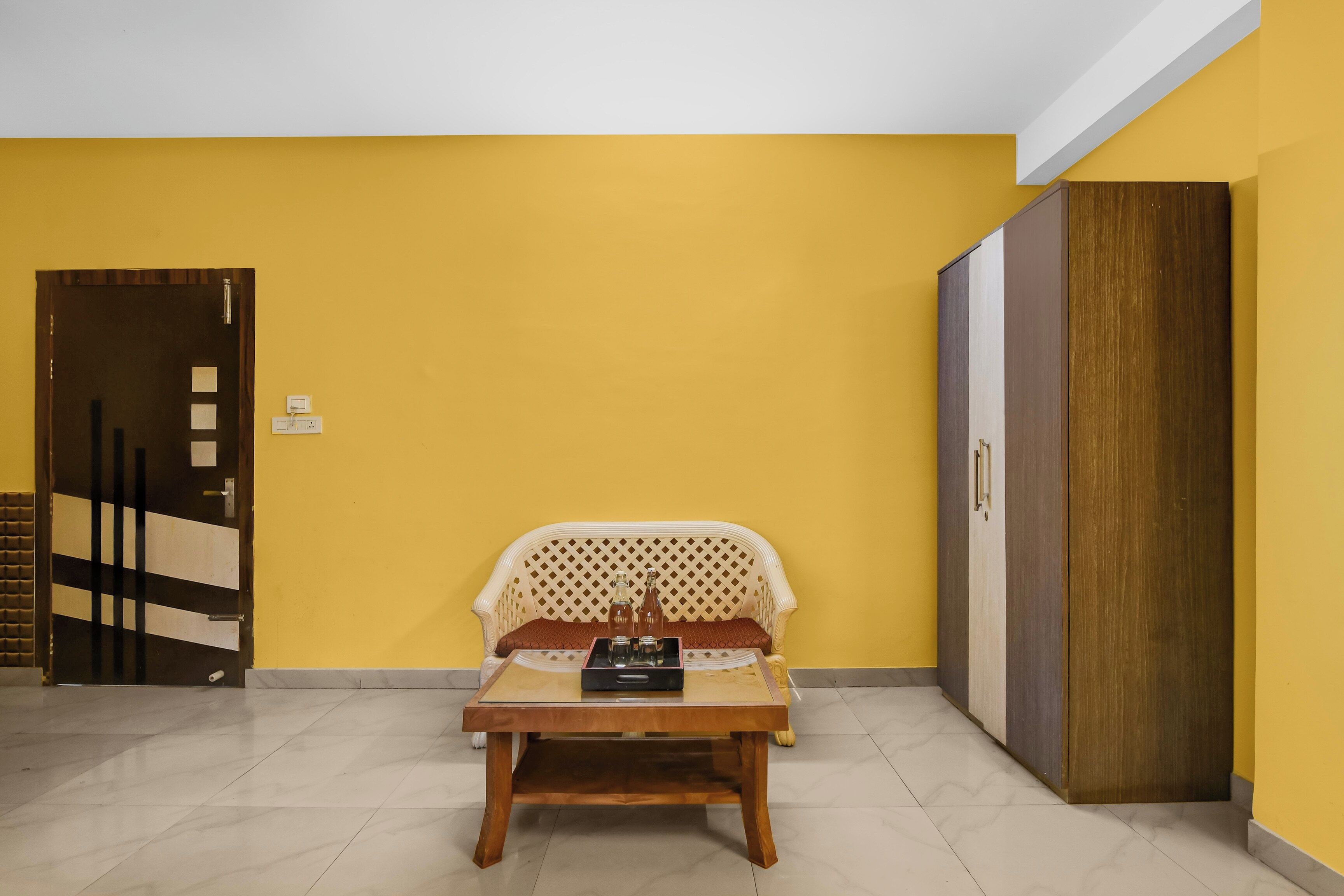 Una habitación con una pared amarilla, una puerta de madera, una silla tapizada blanca con respaldo estampado y una pequeña mesa de madera con una bandeja.