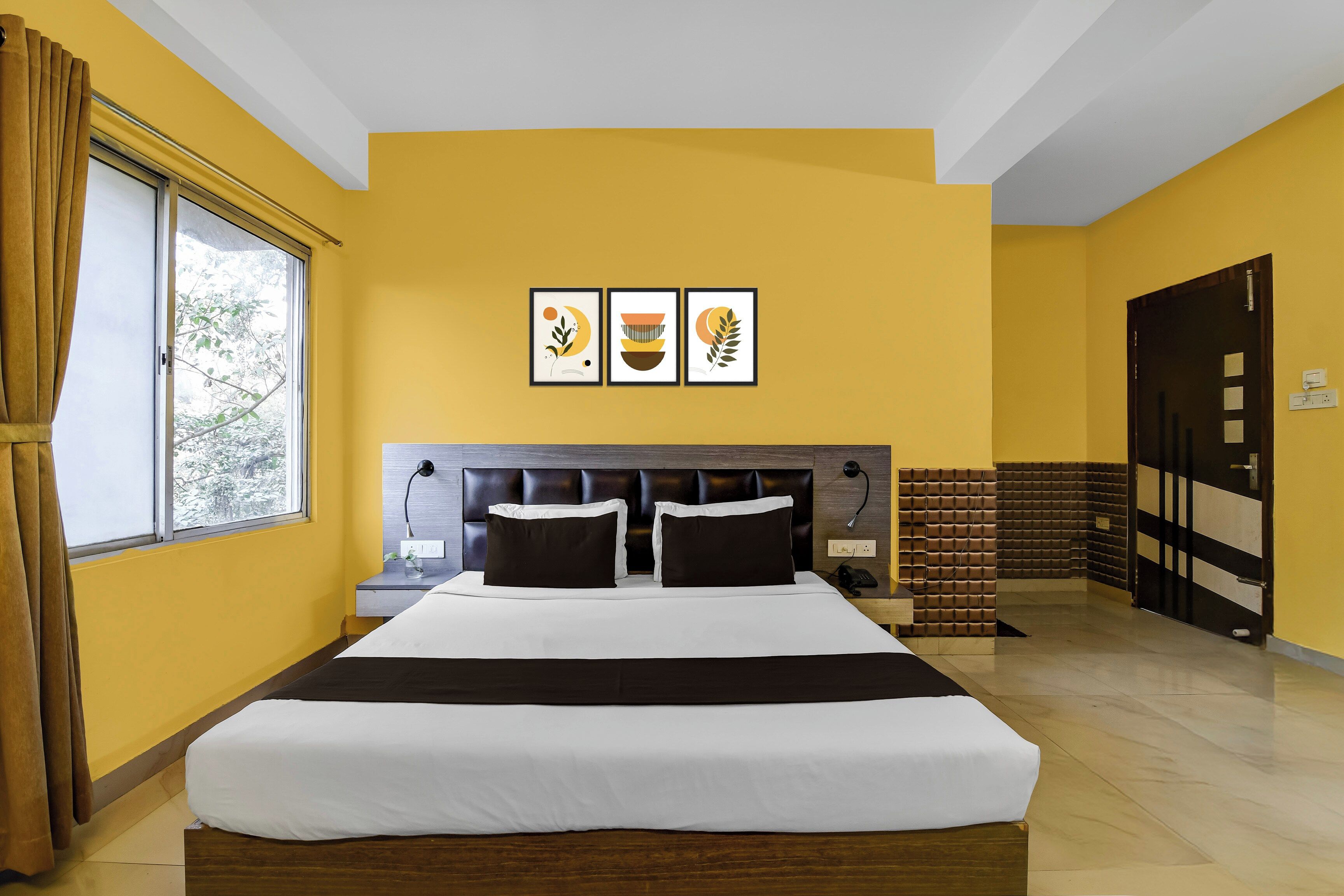 A hotel room with a large bed, a bedside table, a lamp, and a window with curtains. There are three framed pictures on the wall above the bed.