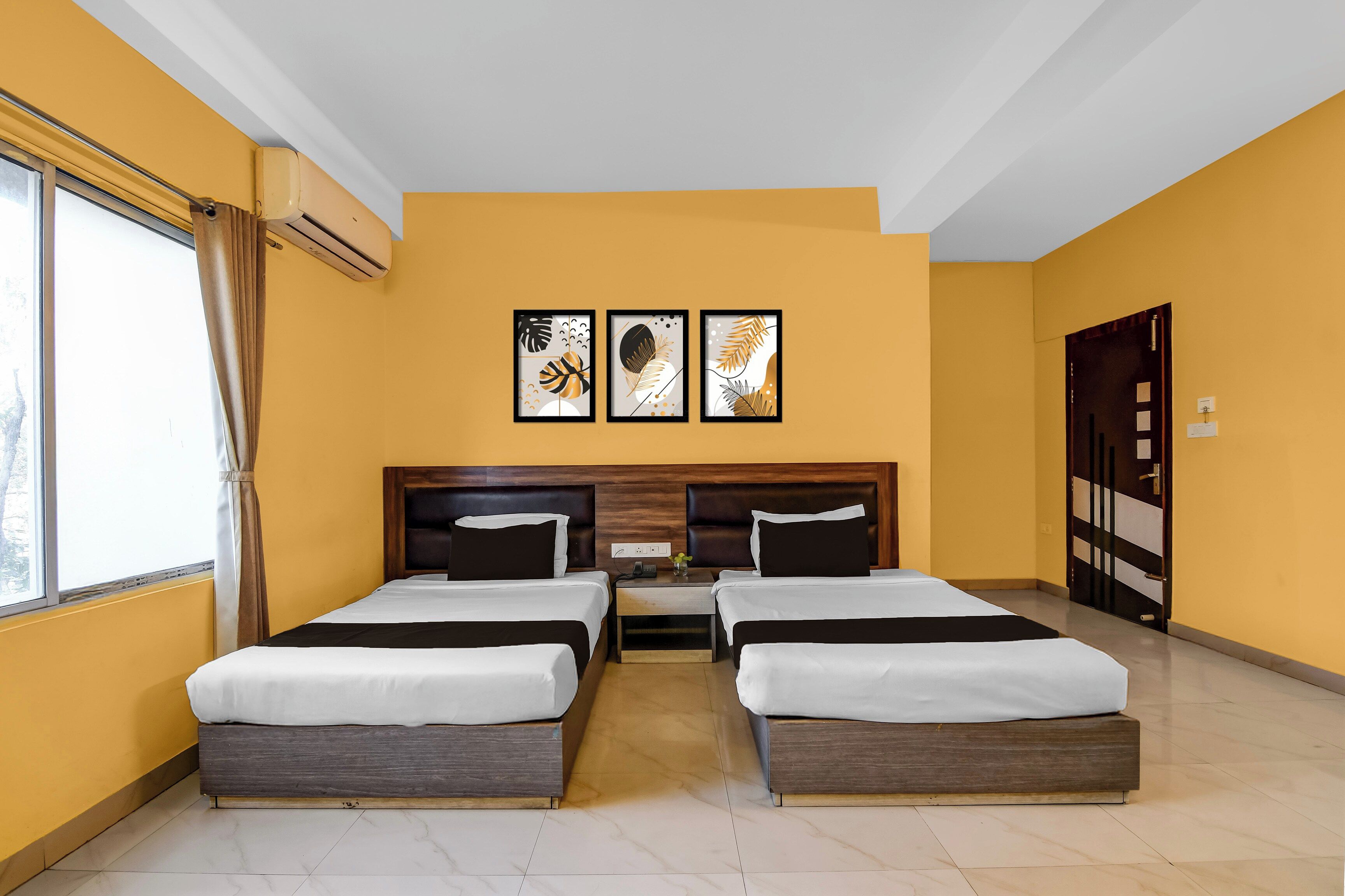 A hotel room with two beds, a window with curtains, and three framed artworks on the wall.