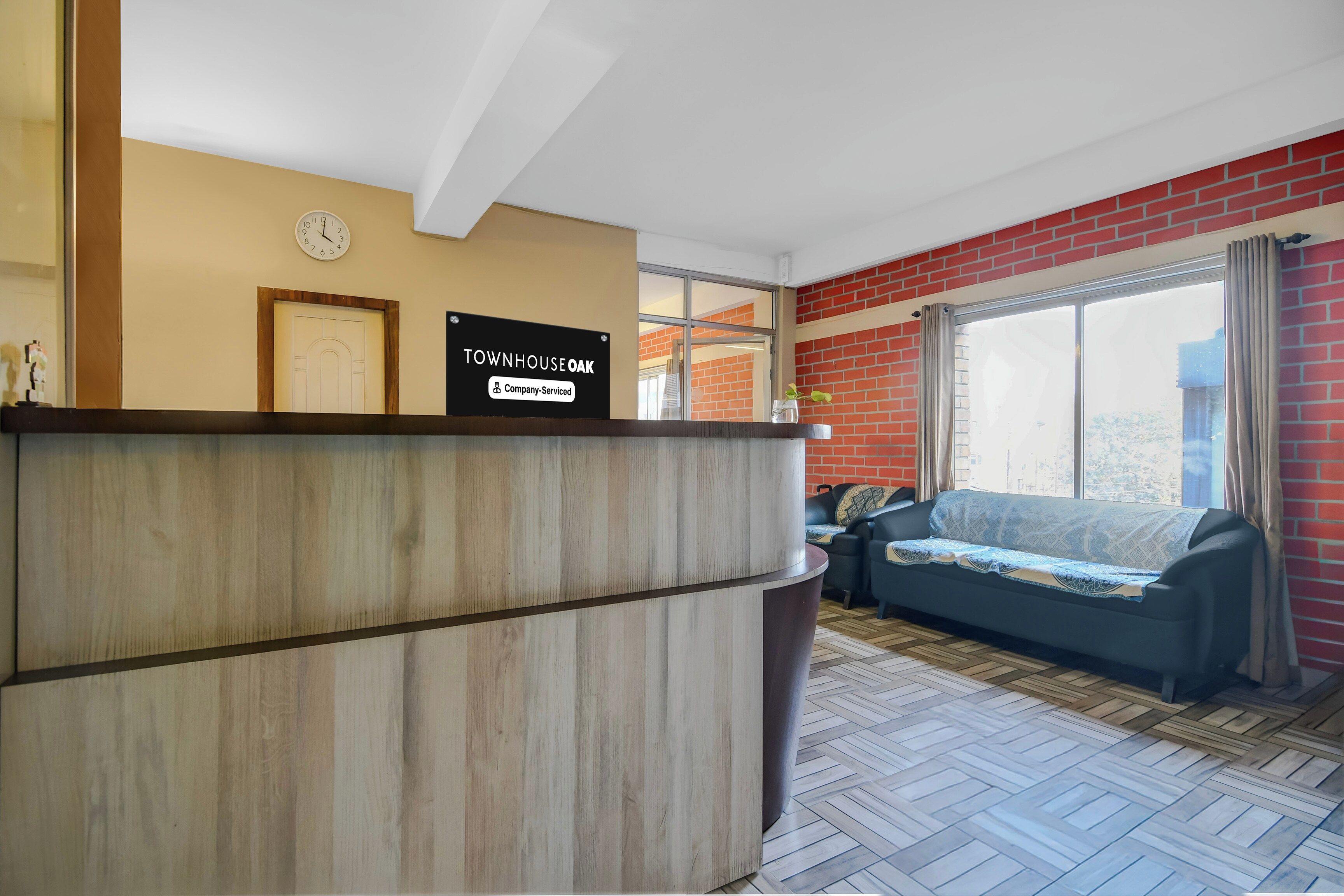 A reception area with a wooden reception desk, a clock, and a sign indicating the property is company-serviced. There is a sofa and a window with curtains in the background.