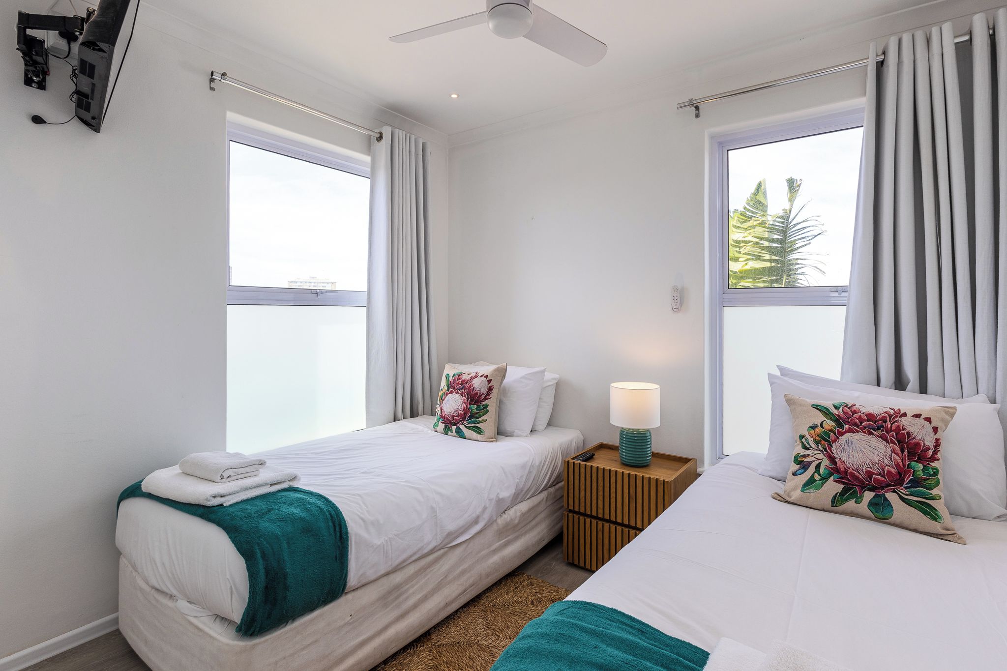 A hotel room with two beds, a ceiling fan, a nightstand with a lamp, and windows with curtains.