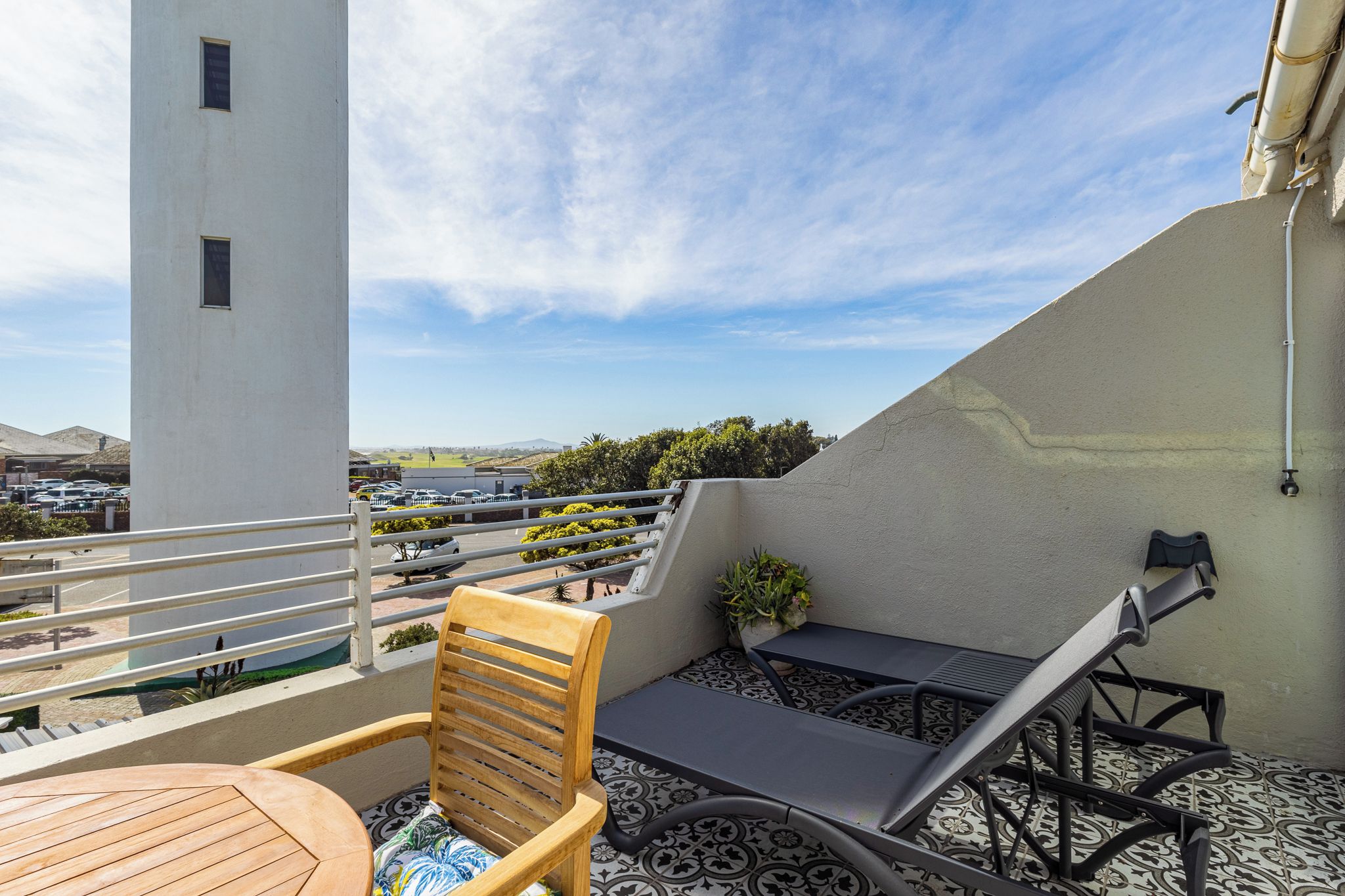 A rooftop terrace with a table, chairs, a lounge chair, and a view of a parking lot and distant landscape.