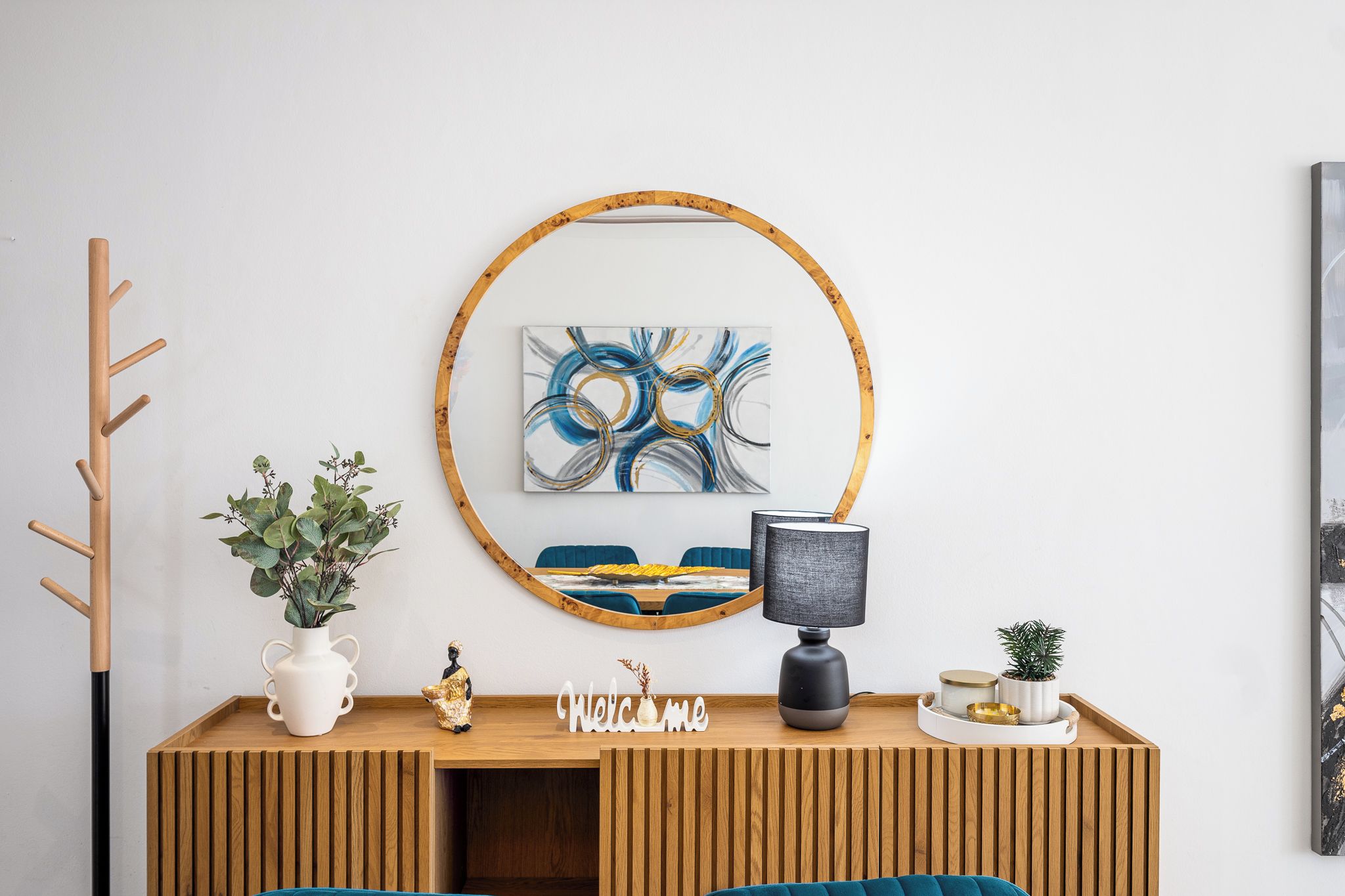 Un salon moderne avec une console en bois, un miroir rond, un tableau, une lampe et un petit panneau d’accueil.